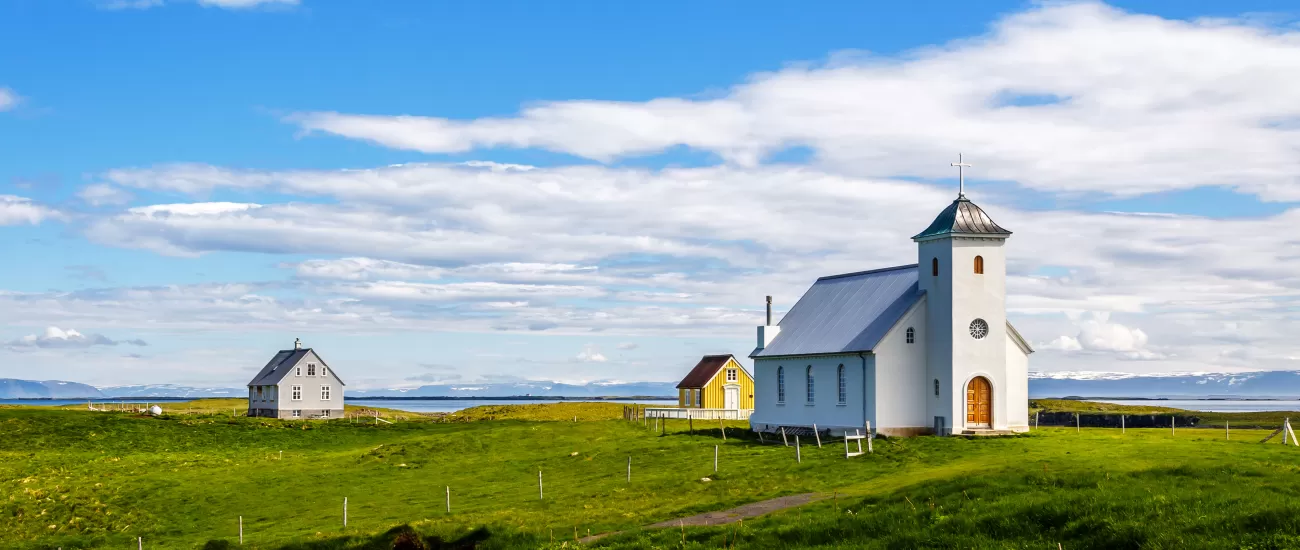 Flateyjarkirkja church and couple of living houses with meadow in foreground and blue sky, Flatey, Iceland