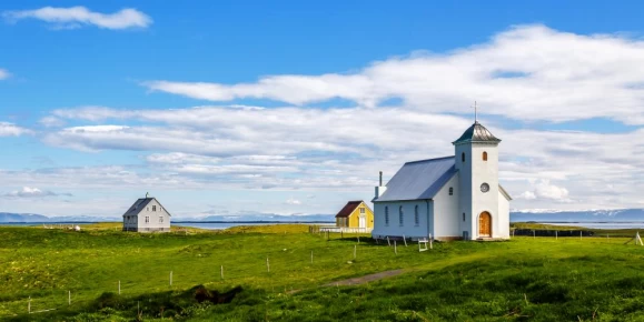 Flateyjarkirkja church and couple of living houses with meadow in foreground and blue sky, Flatey, Iceland