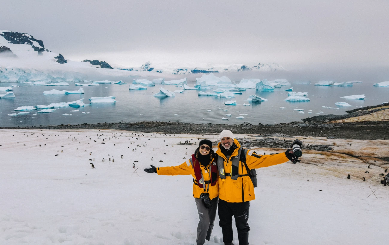 Oksana and Max of Drink Tea and Travel on the snowy Antarctic shores with a colony of Gentoo Penguins in Antarctica.