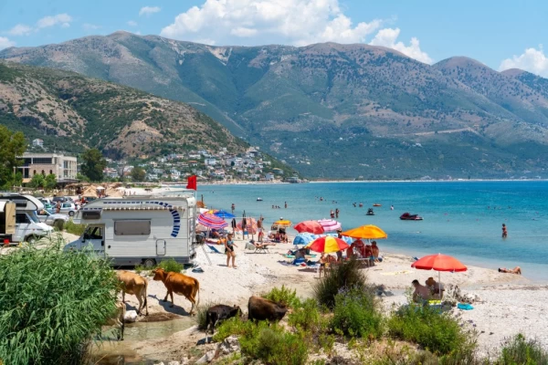 A scenic beach in Albania bustles with holidaymakers under colorful umbrellas, framed by verdant slopes and clear blue skies