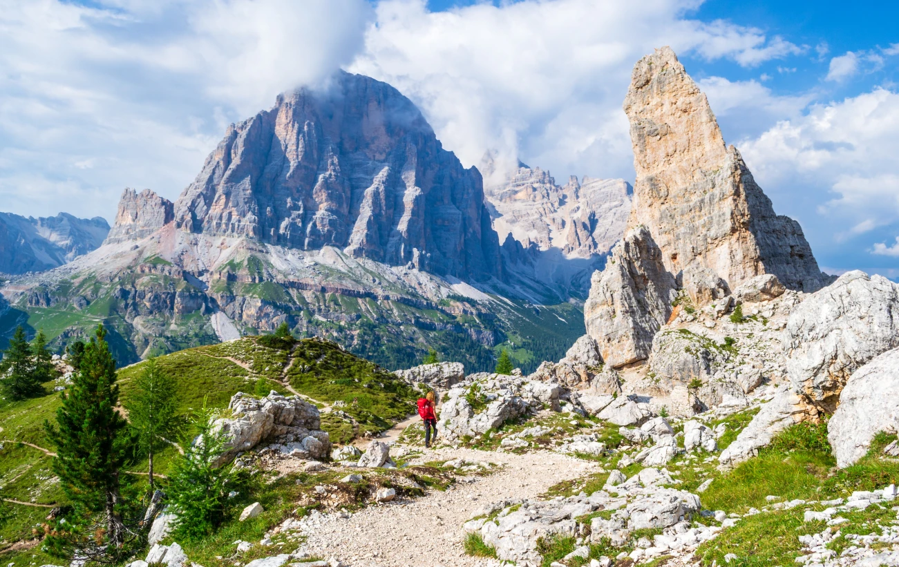 Girl with red backpack on Cinque Torri tour, with views towards Castelletto, Tofana di Rozes, Tofana di Mezzo in the background. Hiking holiday in the Dolomite mountains, Italy.
