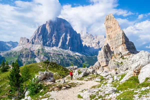 Girl with red backpack on Cinque Torri tour, with views towards Castelletto, Tofana di Rozes, Tofana di Mezzo in the background. Hiking holiday in the Dolomite mountains, Italy.